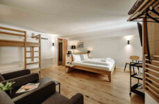 Modernes Hotelzimmer mit Doppelbett, Etagenbett, Sessel und Tisch auf Holzfußboden. Helle Wände und dezente Beleuchtung sorgen für eine einladende Atmosphäre.