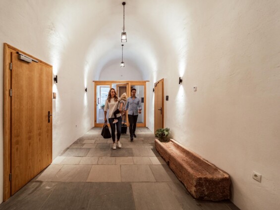 People walking down a well-lit hallway with wooden doors and stone flooring, creating a welcoming atmosphere.