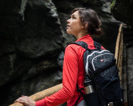 Frau mit rotem Pullover und Rucksack auf einem Holzweg in einer Felsenschlucht.