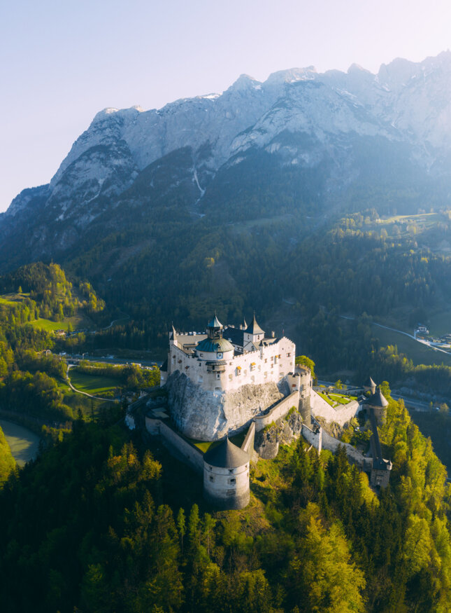 Festung Hohenwerfen vor Alpenpanorama, umgeben von grünen Wäldern bei Sonnenaufgang.