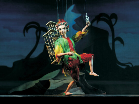 A colorful marionette puppet with a bird-like costume is suspended on stage against a dark, whimsical backdrop.