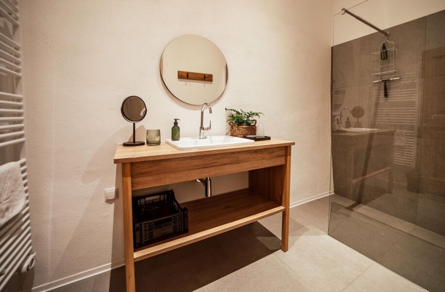 Modernes Badezimmer mit Holzwaschtisch, rundem Spiegel, Glasdusche und minimalistischer Dekoration.