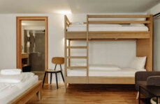 Zimmer mit Etagenbett, Holzstuhl und modernem Bad im Hintergrund.