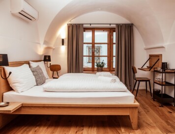 Großzügiges Hotelzimmer mit Doppelbett, Holzboden und Fenster mit Vorhängen. Moderne Einrichtung mit Schreibtisch und Getränkewagen.