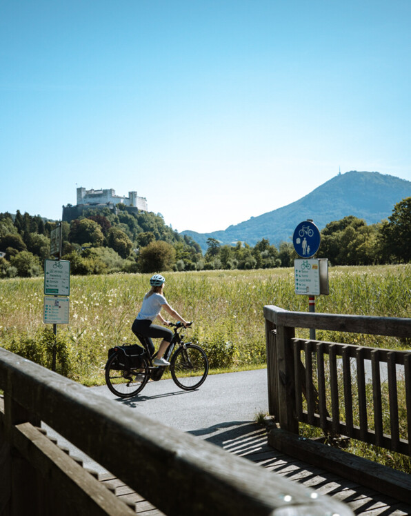 Eine Person fährt mit dem Fahrrad auf einem Weg durch eine grüne Landschaft, mit der Festung Hohensalzburg und einem Hügel im Hintergrund.