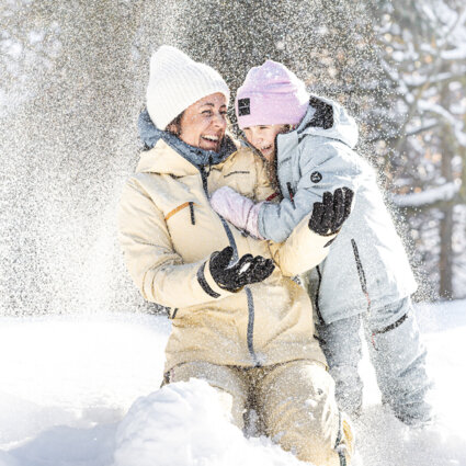Frau mit Kind spielt lachend im Schnee, umgeben von winterlicher Landschaft.