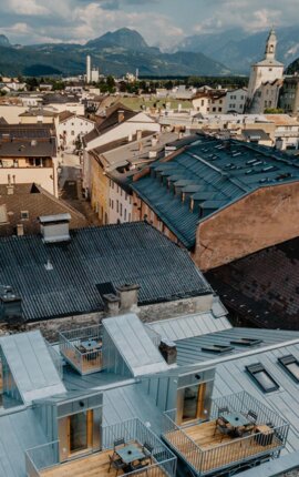 Luftaufnahme von den Rooftop-Apartments mit Blick über die Dächer von Hallein mit Bergen im Hintergrund. Historische Gebäude und malerische Landschaft unter blauem Himmel.