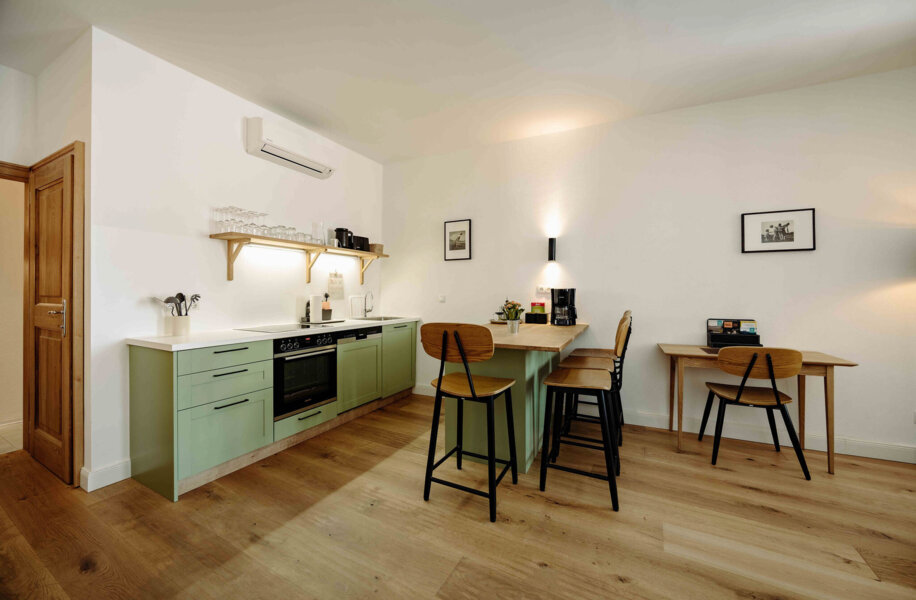 Moderne Küche mit grüner Arbeitsfläche, Barhockern und Esstisch auf Holzboden. Dezentes Licht und minimalistische Dekoration.