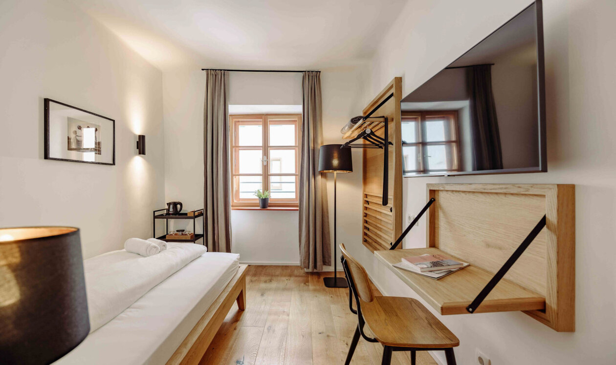 Modernes Hotelzimmer mit Einzelbett, Schreibtisch, Flachbildfernseher und großem Fenster mit Vorhängen, das eine gemütliche Atmosphäre bietet.