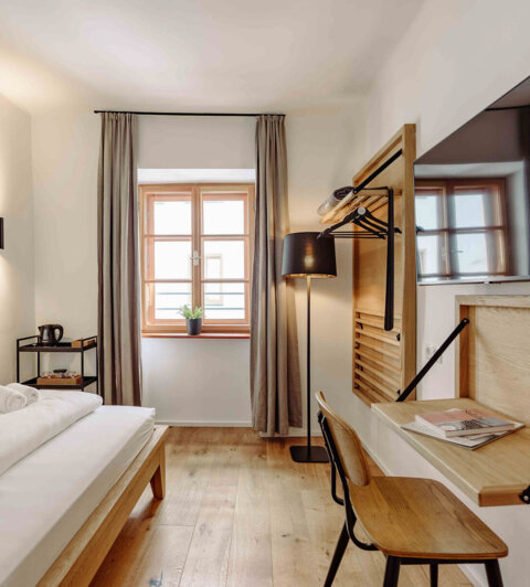 Modernes Hotelzimmer mit Einzelbett, Schreibtisch, Flachbildfernseher und großem Fenster mit Vorhängen, das eine gemütliche Atmosphäre bietet.