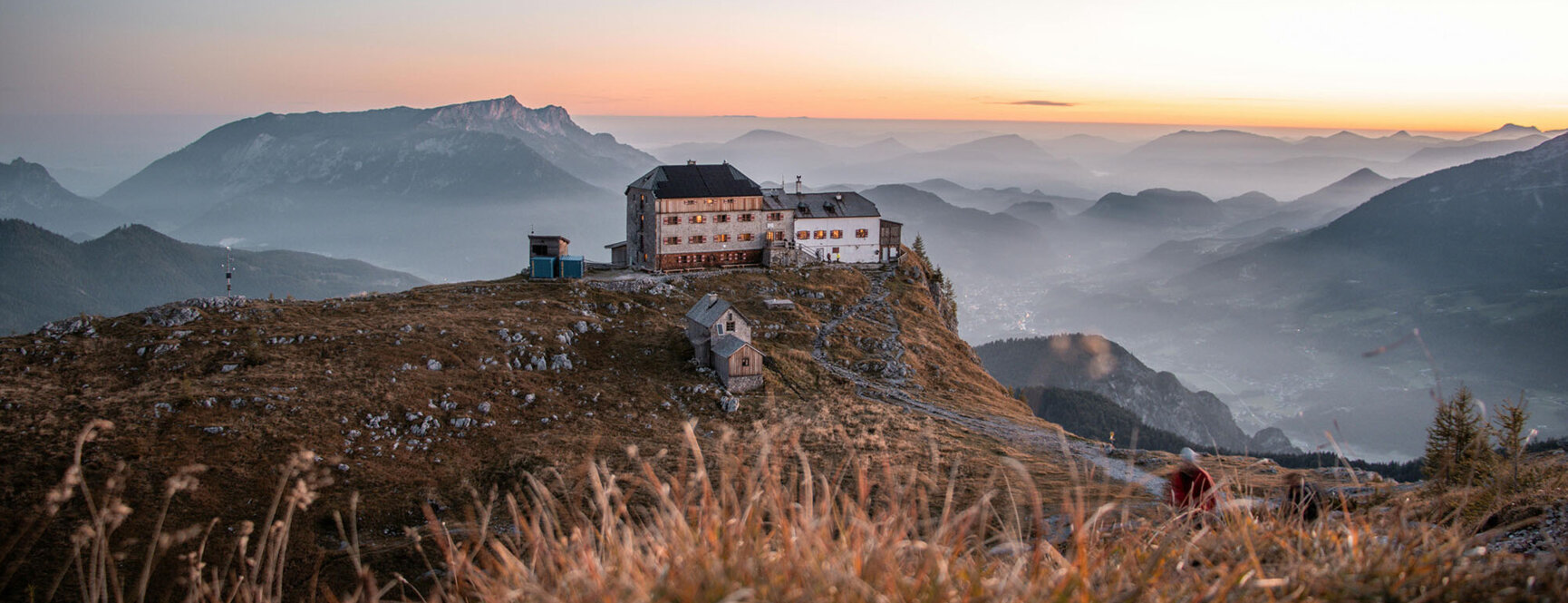 Berglandschaft bei Sonnenaufgang mit einem Gebäude im Vordergrund und Panorama im Hintergrund.