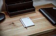 Notizblock mit Stift auf Holzschreibtisch, daneben ein schwarzes Ablagefach und ein geschlossenes Buch.