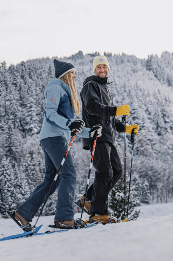 Zwei Personen wandern im Schnee mit Schneeschuhen, umgeben von verschneiten Bäumen und Hügeln, unter einem bewölkten Himmel.
