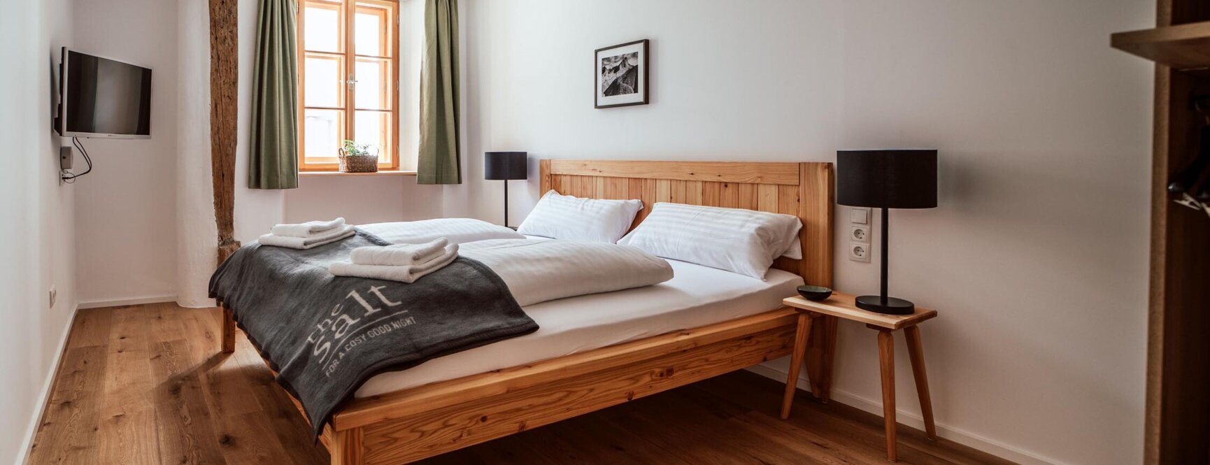 Gemütliches Schlafzimmer mit Holzboden, Doppelbett, TV und Fenster mit grünen Vorhängen in einer modernen Unterkunft.