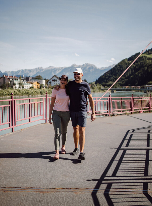 Zwei Menschen spazieren Arm in Arm über eine Brücke in Hallein, mit Blick auf die Salzach und die umliegenden Berge bei Sonnenschein.