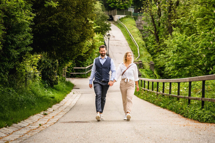 Ein Paar spaziert auf einem Weg vor der Festung Hohensalzburg, umgeben von üppigem Grün.