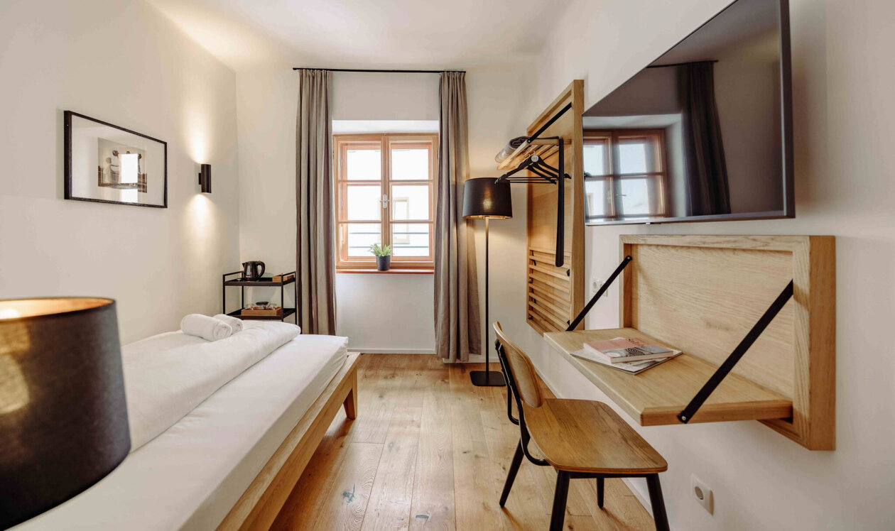 Modern hotel room with a single bed, wooden floors, a wall-mounted TV, a desk, and a window with curtains.