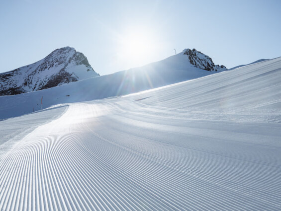 Perfekt präparierte Skipiste bei Sonnenschein, umgeben von verschneiten Bergen.