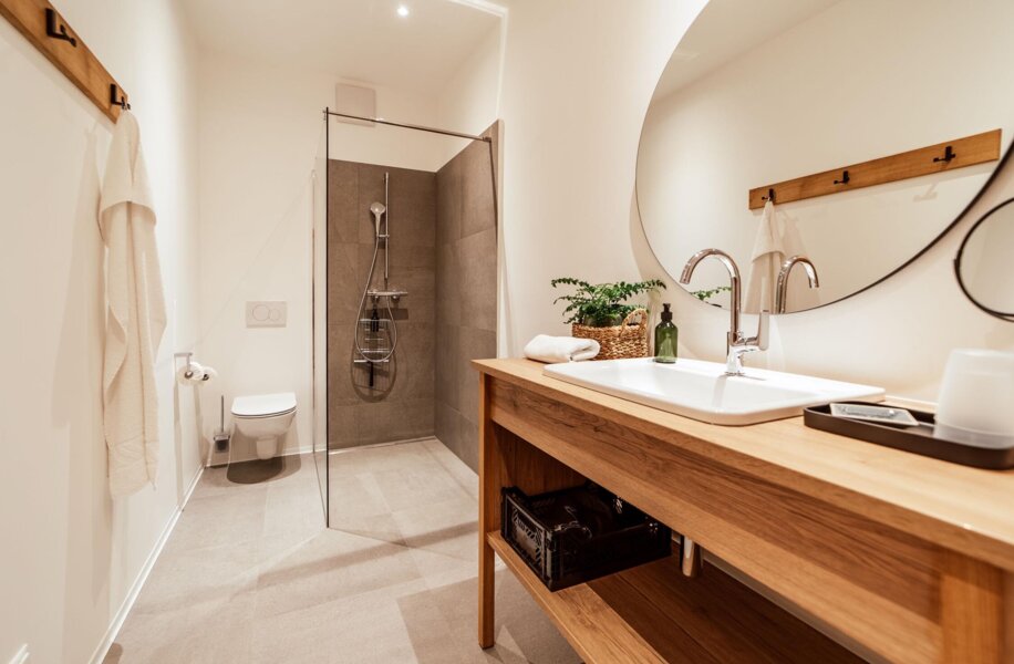 Modern bathroom with a wooden vanity, round mirror, and walk-in shower. Minimalist design featuring neutral colors and natural accents.