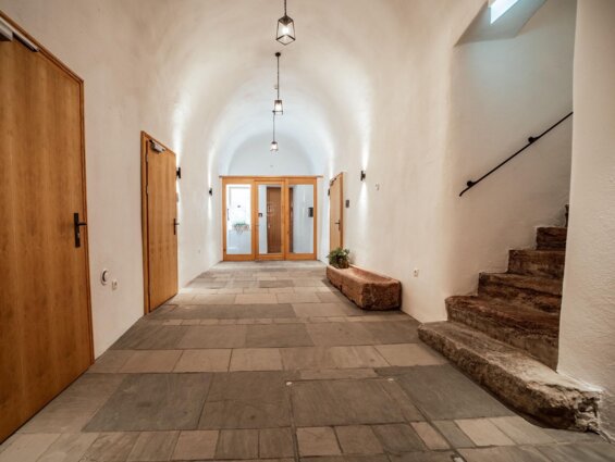Heller Flur mit gewölbter Decke, Holztüren und Steinböden, führt zu Glastüren. Rechts eine Steintreppe mit Geländer. Moderne Beleuchtung.