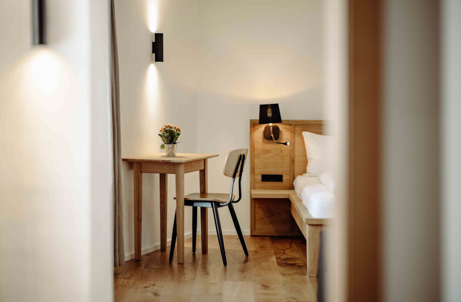 Ein minimalistisches Hotelzimmer mit Holzmöbeln, Tisch, Stuhl und Bett. Eine stilvolle Beleuchtung und Blumen sorgen für eine gemütliche Atmosphäre.