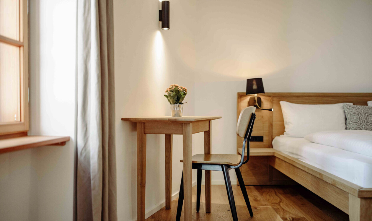 Gemütliches Zimmer mit Holztisch, Stuhl und Bett; natürliche Beleuchtung und helle Einrichtung.