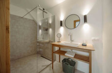 Modernes Badezimmer mit begehbarer Dusche, Holz-Waschtisch und rundem Spiegel. Dezente Beleuchtung betont die schlichte Eleganz des Raumes.