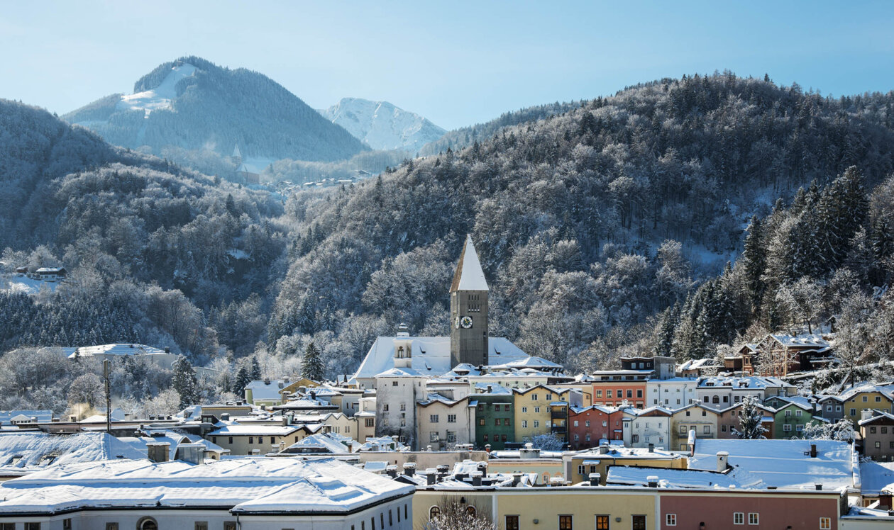 Snow-covered Hallein with colorful buildings and a church tower, nestled in a mountainous landscape under a clear blue sky.