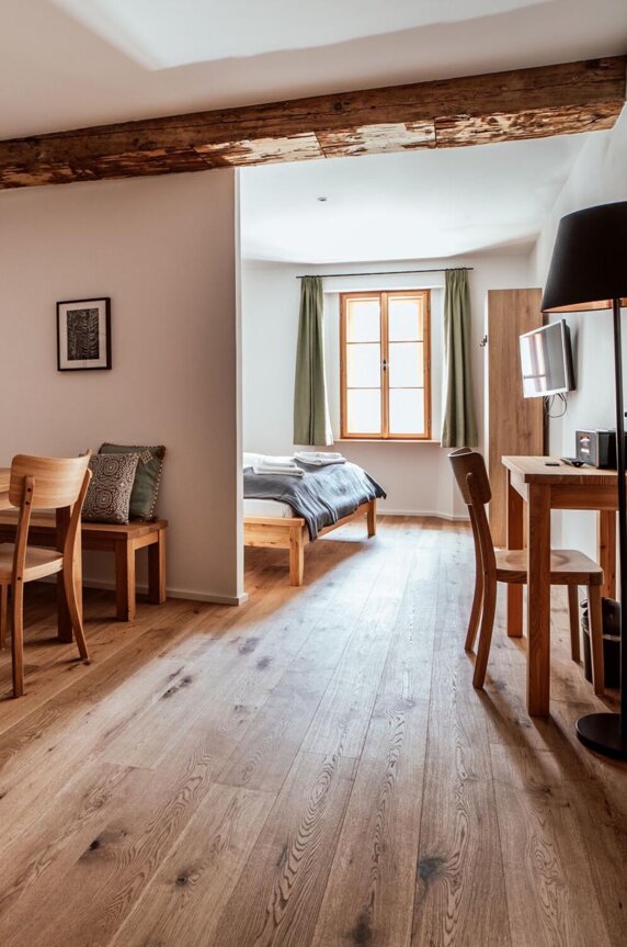 Gemütlicher Wohnbereich mit Holzboden, Esstisch, Stühlen und einem Schreibtisch. Im Hintergrund ist ein Bett mit Fenster zu sehen.