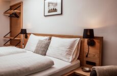 Cozy hotel room with a wooden bed, white linens, decorative pillow, and two wall lamps. Minimalist design with open shelving and a framed photo.