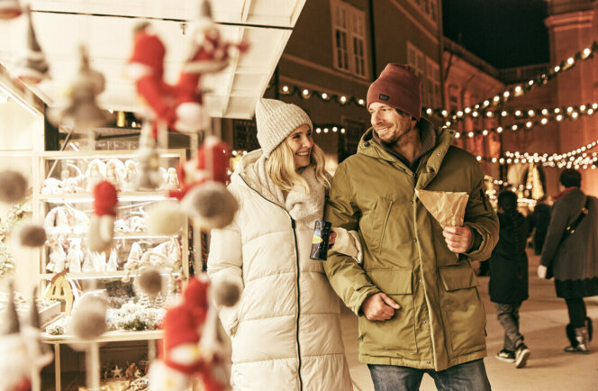 Ein Paar spaziert über einen stimmungsvollen Weihnachtsmarkt in Hallein und genießt Punsch und Maroni unter Lichterketten.