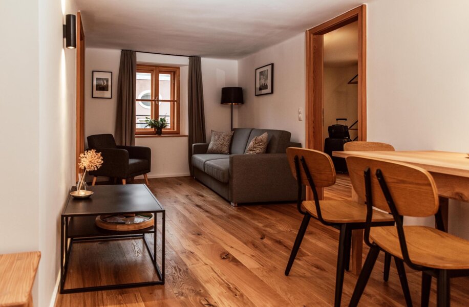 Cozy living area with wooden flooring, a grey sofa, armchair, and dining table, featuring warm lighting and a window with curtains.
