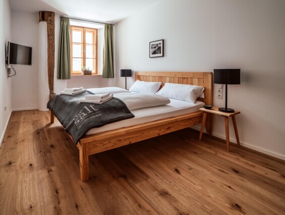 Gemütliches Schlafzimmer mit Holzboden, Doppelbett, TV und Fenster mit grünen Vorhängen in einer modernen Unterkunft.