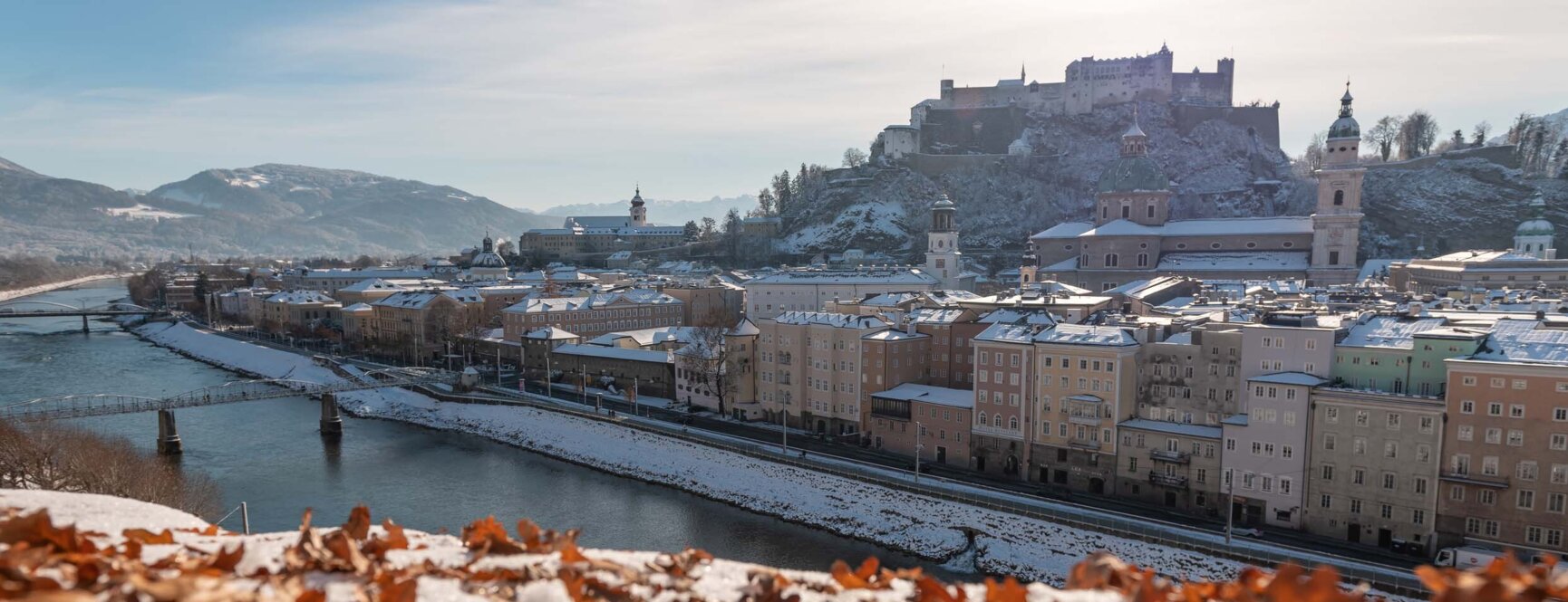 Winterlicher Blick über die Salzach auf Salzburgs Altstadt mit schneebedeckten Dächern und der Festung Hohensalzburg.