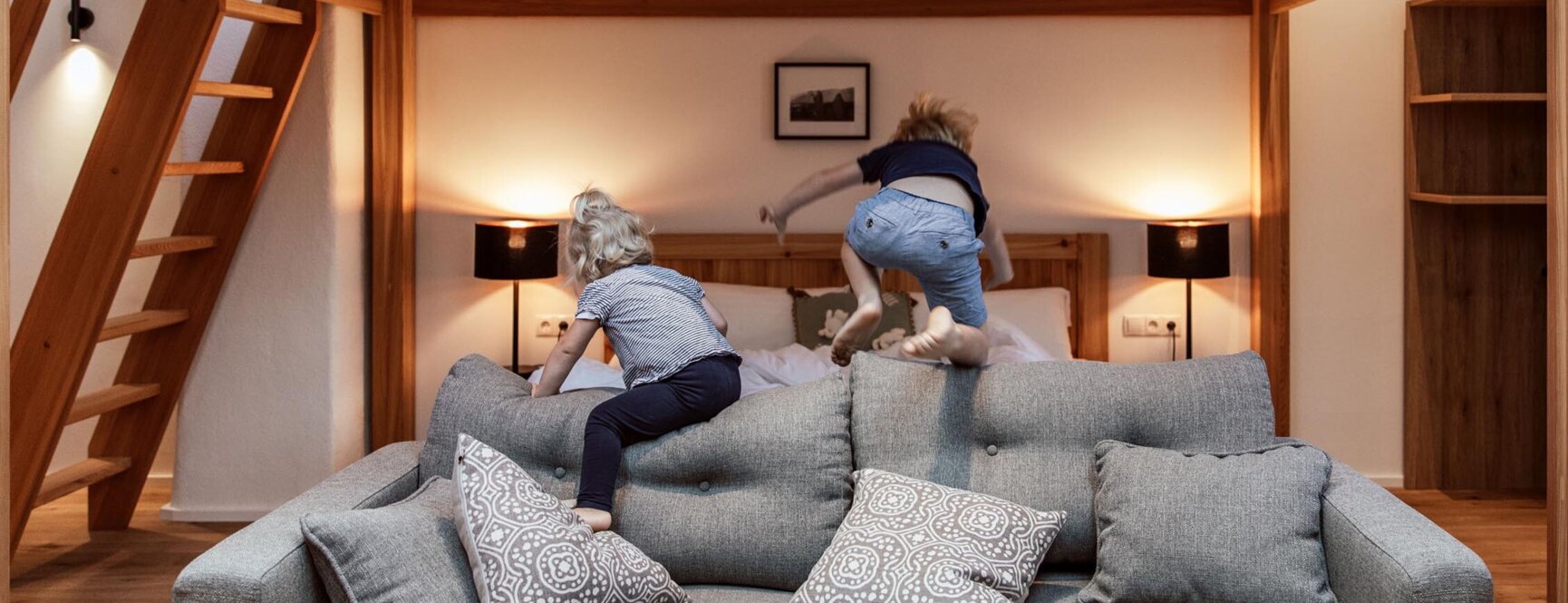 Children playing on a bed in a cozy, wooden-furnished loft room, featuring a gray sofa and decorative pillows.