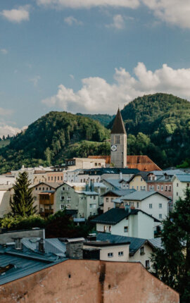 A picturesque view of Hallein with colorful buildings, church towers, and lush green hills under a partly cloudy sky.