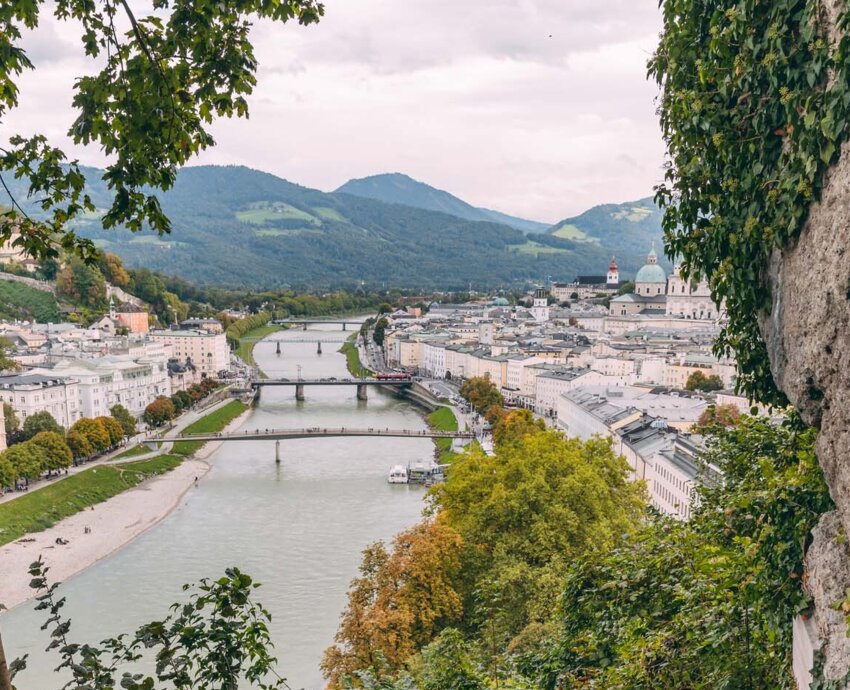 Zwei Personen steigen eine bewaldete Treppe hinauf, mit Blick über Salzburg, die Salzach und ihre Brücken im Hintergrund.