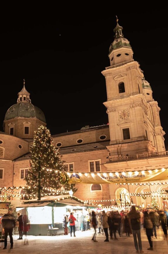 Weihnachtsmarkt in Salzburg bei Nacht, beleuchtet mit Lichtern und einem großen Weihnachtsbaum, vor einer historischen Kirche, gemütliche Atmosphäre.