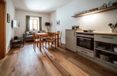 Wohnbereich mit offener Küche, Esstisch und grauem Sofa, durch ein Fenster kommt Tageslicht herein.