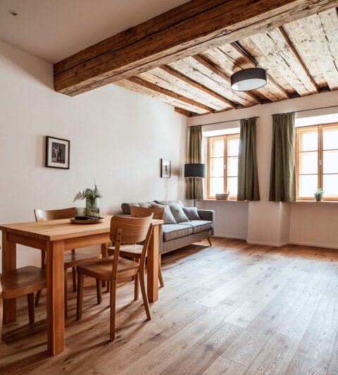 Cozy living room with wooden beams, a dining table, and a sofa. Large windows with green curtains bring in natural light.