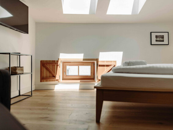 Helles Hotelzimmer mit Dachfenster, Bett, Fernseher und Regal, Sonnenschein dringt durch die Fenster.