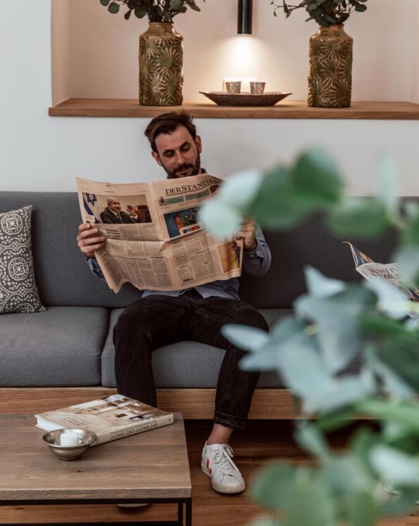 Man reading a newspaper on a cozy sofa, with decorative plants and a coffee table in a stylish room.