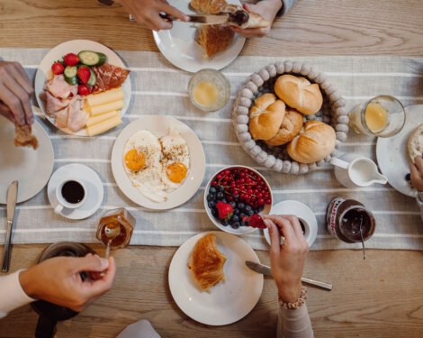 Hands sharing a fresh breakfast spread: rolls, croissants, eggs, cold cuts, cheese, berries, coffee, and juice on a striped table runner.