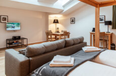 Gemütlicher Wohnraum mit Sofa, TV, Esstisch und Schreibtisch, im Hintergrund ein Doppelbett