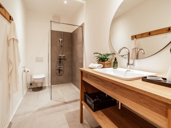 Modernes Badezimmer mit begehbarer Dusche, hölzernem Waschtisch, großem Spiegel und dekorativer Pflanze. Minimalistisches, helles Design.