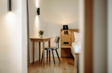 Ein minimalistisches Hotelzimmer mit Holzmöbeln, Tisch, Stuhl und Bett. Eine stilvolle Beleuchtung und Blumen sorgen für eine gemütliche Atmosphäre.