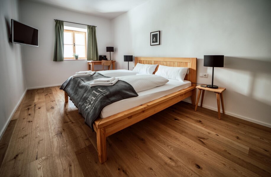 Großzügiges Schlafzimmer mit Holzboden, Doppelbett, TV und natürlichem Licht durch ein Fenster.