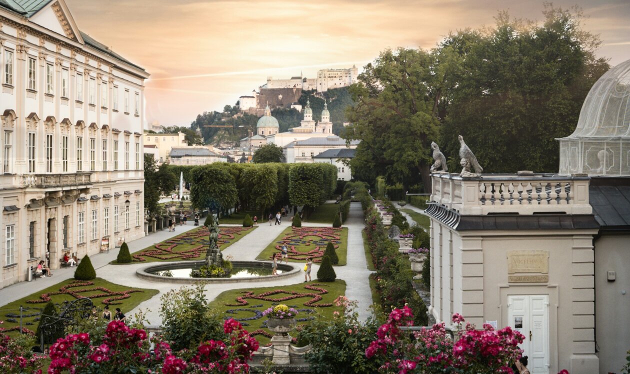 Three people sit on grass near vibrant rose bushes, overlooking Mirabell palace and garden and the historic fortress under a dramatic sky at sunset.
