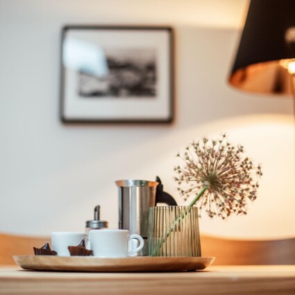 Tablett mit Kaffeetassen, Zuckerdose und dekorativer Blume auf einem Tisch, im Hintergrund eine Lampe und ein Bild an der Wand.