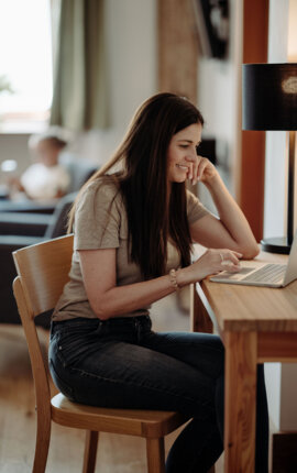 Frau arbeitet lächelnd am Laptop an einem Holztisch in einem gemütlichen Apartment in Hallein.